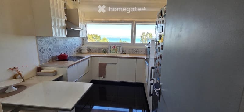 Bright kitchen with white cabinets, black tiled floor, and a large window offering a sea view. Includes a refrigerator, sink, oven, countertop, and various kitchen items.