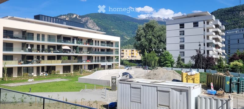 Modern multi-story residential building with balconies and large windows, surrounded by greenery and mountains in the background.