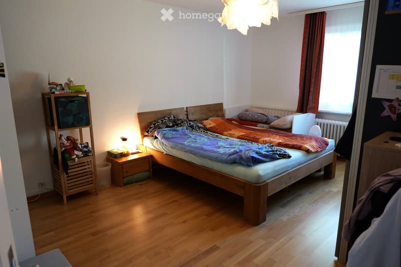 Cozy bedroom with wooden flooring, a double bed with colorful bedding, a nightstand with a lamp, a wooden shelf with toys and storage boxes, and a window with curtains.