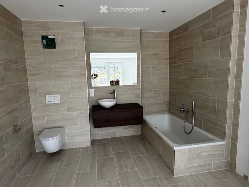 Modern bathroom with a bathtub, toilet, sink, and mirror. The walls and floor are tiled in a neutral tone, and the sink is mounted on a dark wooden vanity.