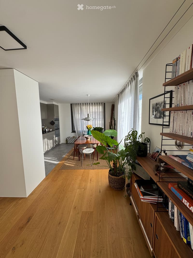 Bright, open living space with wooden flooring, large windows, bookshelf, dining area, and kitchen visible in the background.