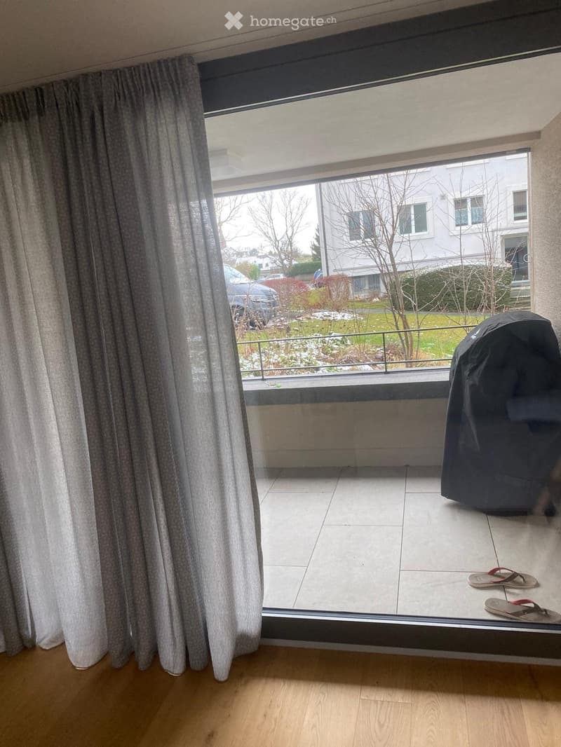 Balcony with tiled floor, large window with curtains, view of garden and neighboring building, flip-flops placed on floor.