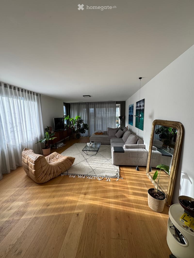 Modern living room with wooden flooring, large windows, a grey sectional sofa, a glass coffee table, a beige armchair, a large mirror, and several potted plants.