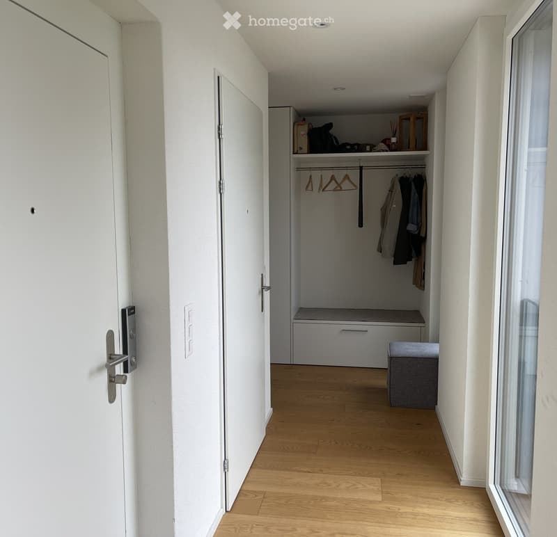 A modern hallway with light wooden flooring, white walls, and a built-in closet with hanging space and a bench. A small grey ottoman is placed near the window.