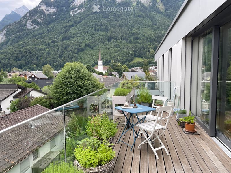 Balcony with wooden decking, glass railing, small table and chairs, potted plants, overlooking a town and mountain landscape.