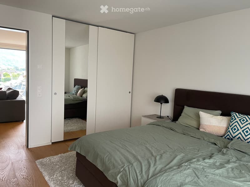 Modern bedroom with a large bed, nightstand, lamp, and a mirrored closet. The room features light-colored walls, wooden flooring, and a view of an outdoor area through an open door.