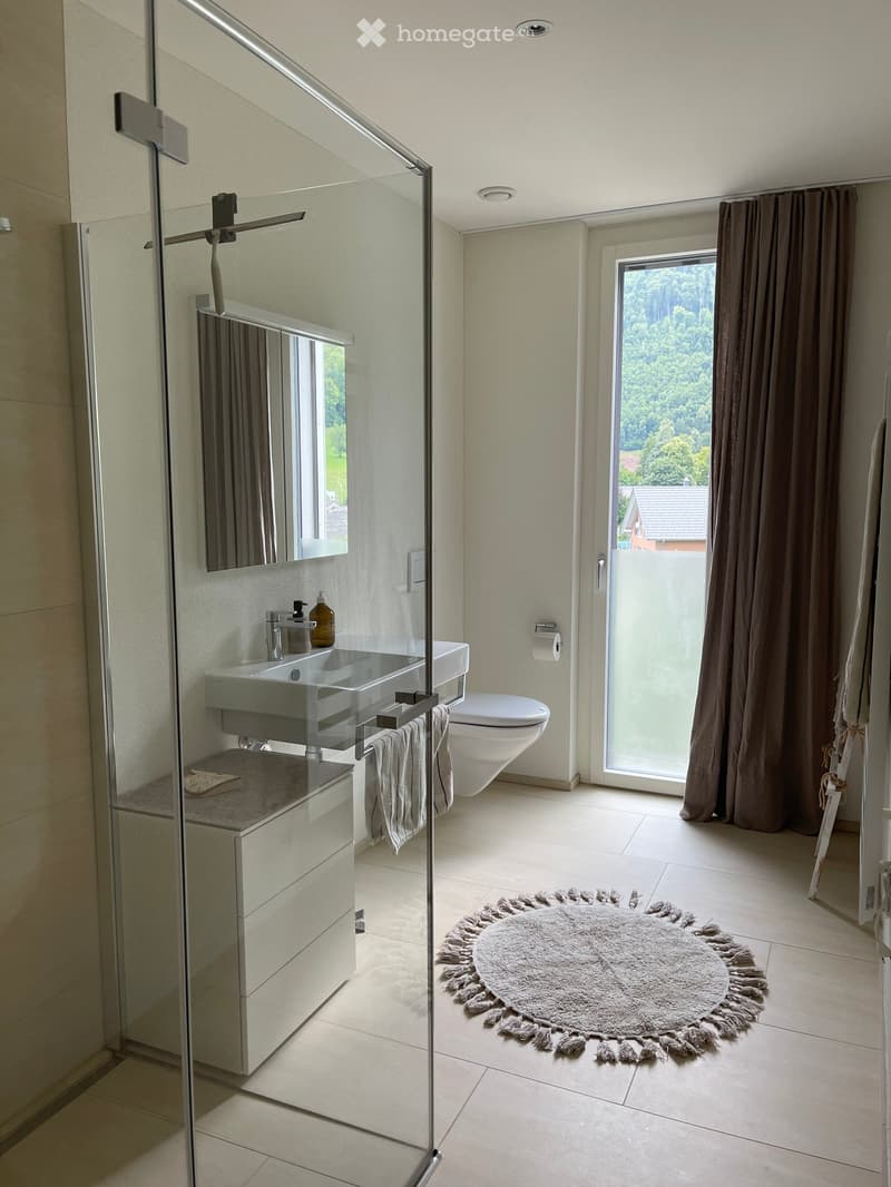 Modern bathroom with a glass-enclosed shower, white sink, toilet, and a large window with a view of greenery. Includes a round rug, towels, and a mirror.