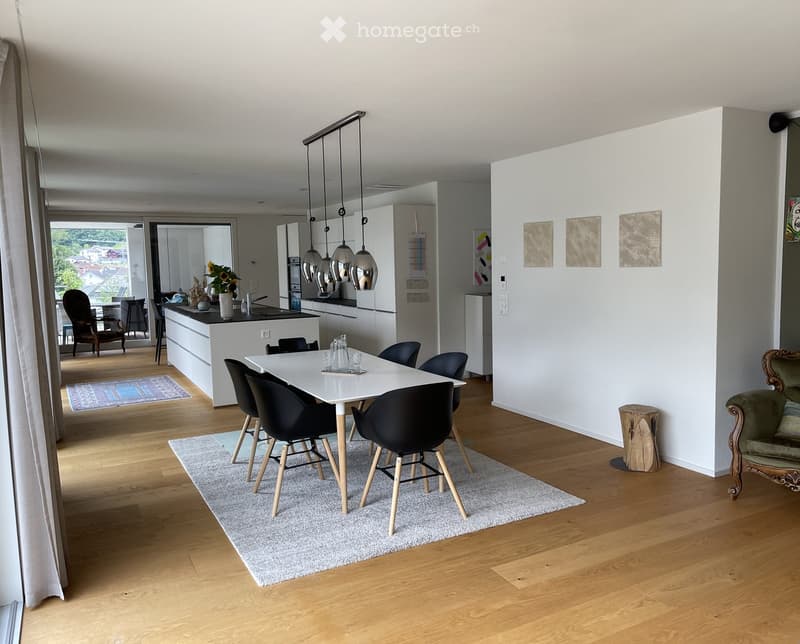 Modern open-plan living and dining area with light wood flooring, white dining table with black chairs, grey sofa, kitchen island with pendant lights, and a view through large windows.