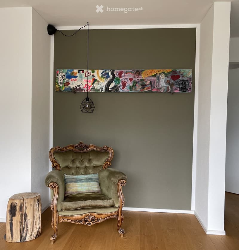 A small, elegantly decorated room with a tufted green sofa, a wooden stool, a colorful abstract painting on the wall, and a modern pendant light hanging from the ceiling.