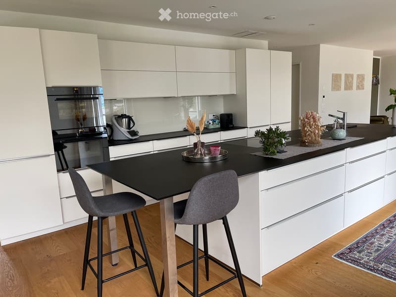 Modern kitchen with white cabinets, black countertops, and a central island with two stools. Includes an oven, coffee maker, and decorative items on the counters.