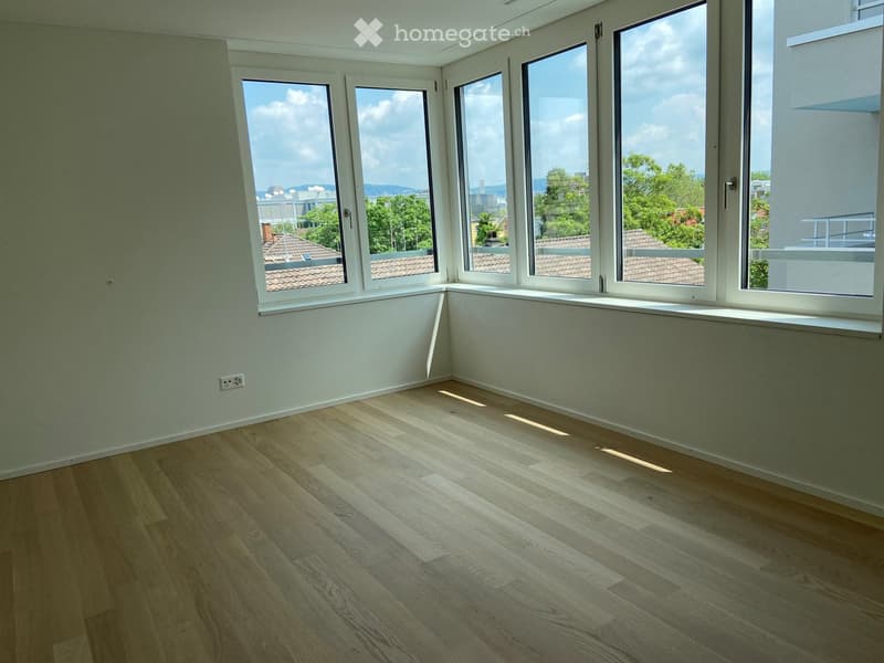 Empty room with light wooden flooring and large windows offering a view of rooftops and trees.