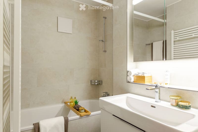 Modern bathroom with a bathtub, sink, and large mirror. Includes a towel rack, decorative tray with candles and a small plant, and a wooden shelf with folded towels and toiletries.