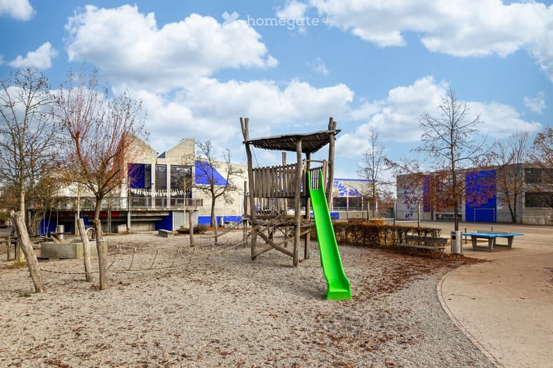 Outdoor playground area with a green slide, wooden climbing structure, benches, and a ping pong table, surrounded by trees and modern buildings.
