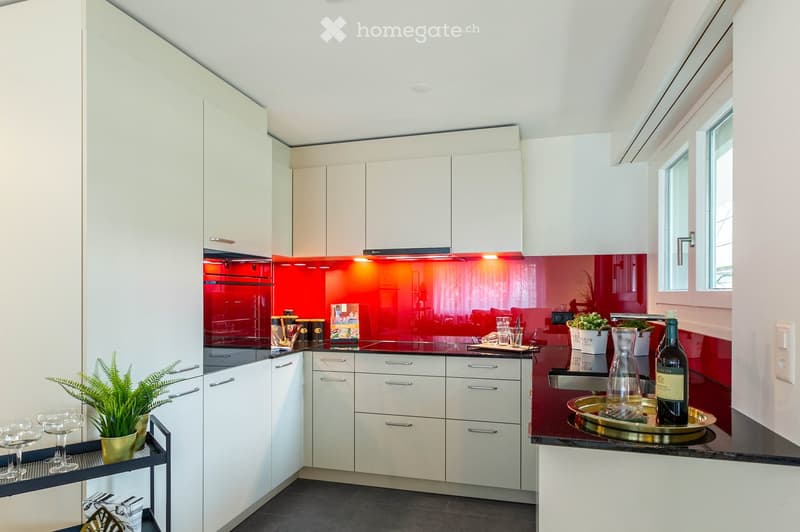 Modern kitchen with white cabinets, red backsplash, and stainless steel appliances. Features include a countertop with a bottle, vase, potted plant, and wine glasses.