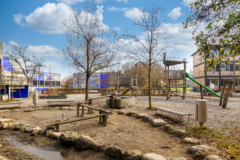 Outdoor playground area with wooden seesaw, slide, benches, and scattered rocks, surrounded by trees and modern buildings.