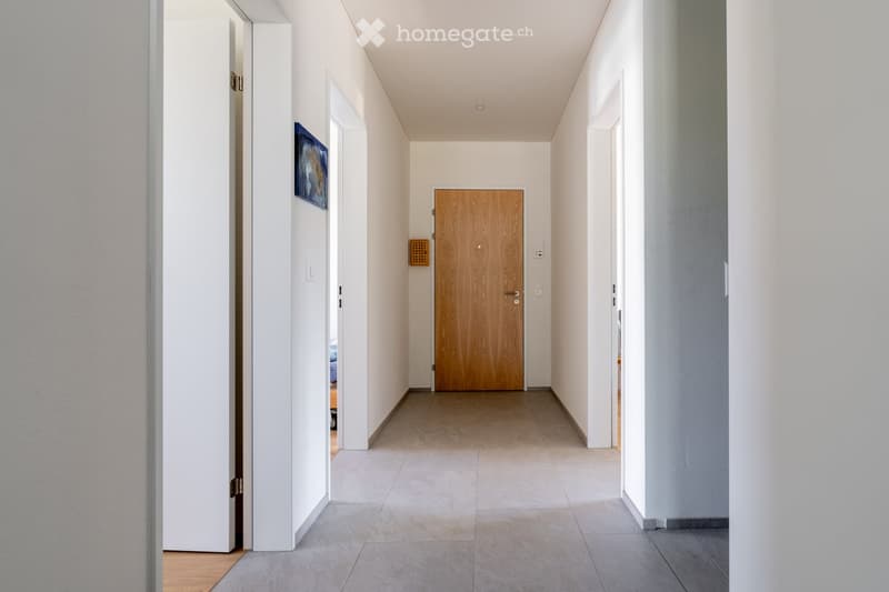 A modern, minimalist hallway with white walls, tiled floor, and a wooden door at the end. Features include recessed ceiling lighting, a small wall-mounted artwork, and a ventilation grille on the left wall.