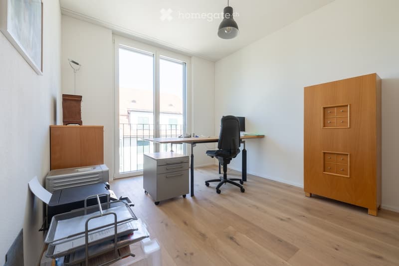 A modern, well-lit office space with a wooden floor, large windows, and a desk setup including a chair, screen, and filing cabinet. Additional furniture includes a wooden cabinet, a printer, and a lamp.