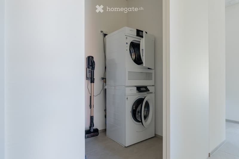 A small, well-lit room with a stacked washing machine and dryer unit, a vacuum cleaner leaning against the wall, and tiled flooring.