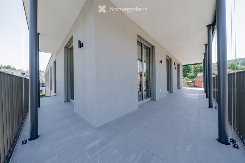 Modern balcony with tiled flooring, glass railings, and metal support columns. The space is open and clean, with a view of the surrounding area.
