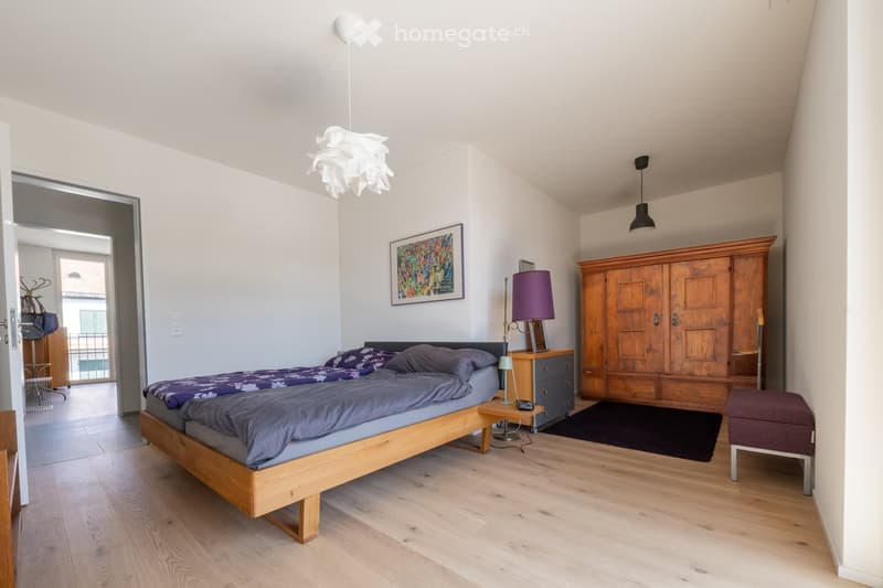 Bright bedroom with wooden flooring, a large bed with purple bedding, a nightstand with a lamp, a wooden wardrobe, and a small stool. A colorful picture frame hangs on the wall, and a balloon is attached to the ceiling.