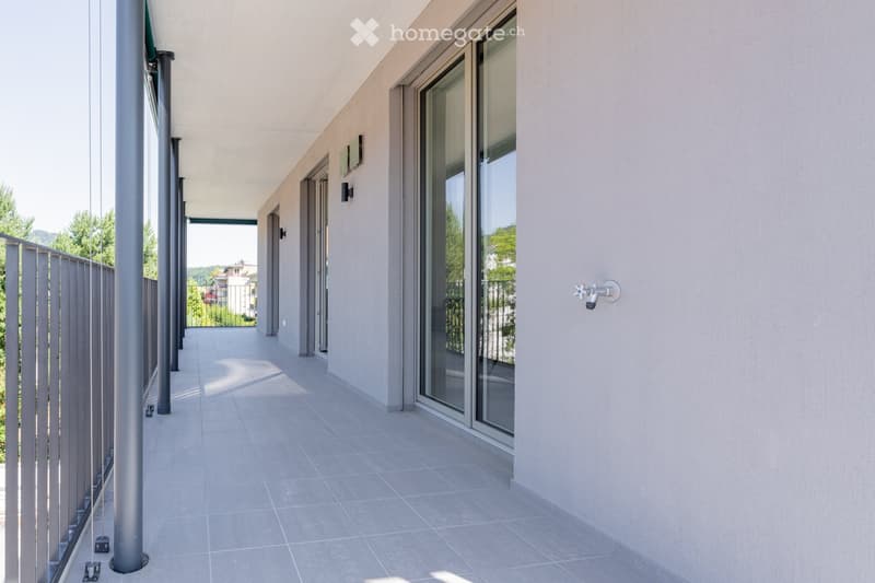 Modern balcony with large glass sliding doors, tiled floor, and metal railing, overlooking a green area with trees and distant buildings.