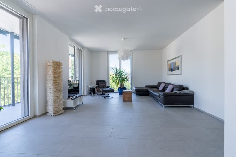 Modern living room with large windows, grey sofa, leather chair, small table, potted plant, and decorative sculpture.