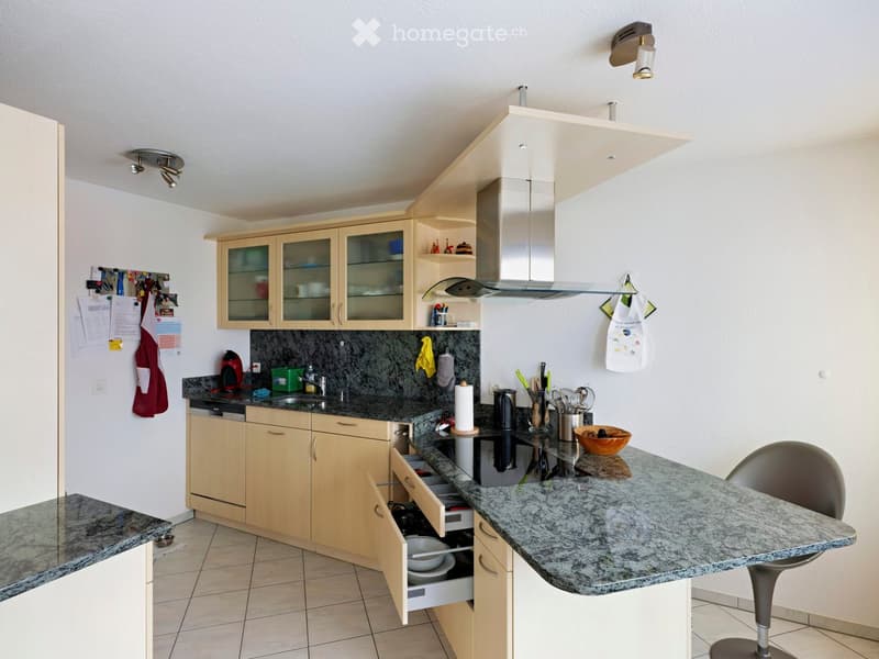 Modern kitchen with light wood cabinets, granite countertops, stainless steel appliances, and a central island with a bar stool.