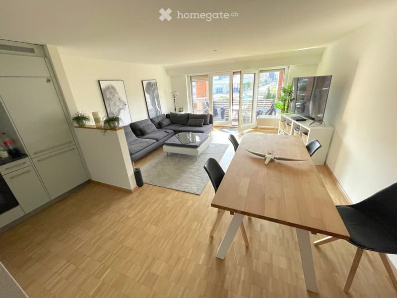 Modern living room with a grey sectional sofa, coffee table, dining table with chairs, TV, and decorative plants.