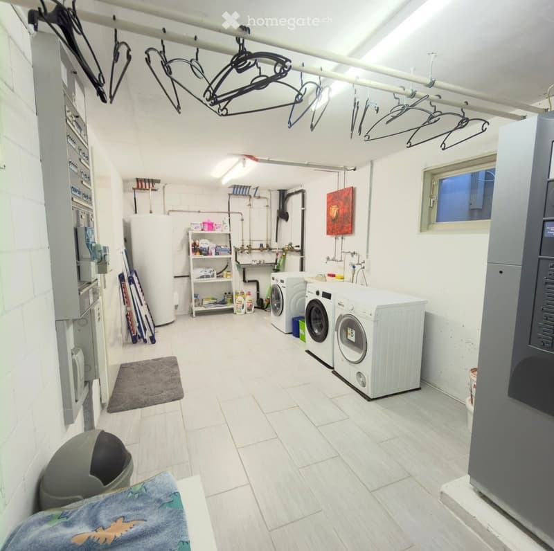 Well-lit laundry room with white tiled floors, white walls, and a window. Includes a washing machine, dryer, shelf with cleaning supplies, and hanging clothes.