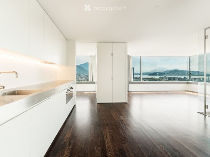 Modern, minimalist kitchen with white cabinets, a dark wooden floor, and expansive windows offering a scenic view of mountains and a cityscape.