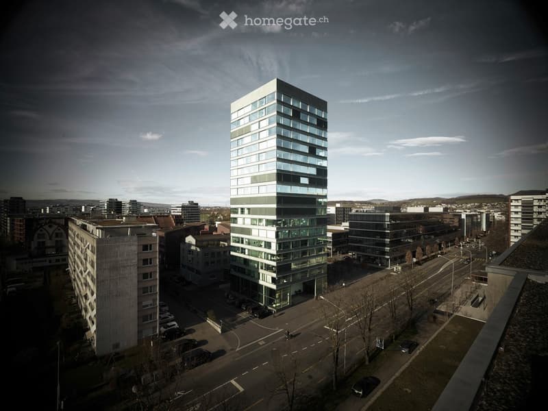 A modern, multi-story building with large glass windows and a sleek design, surrounded by other structures and a parking area.