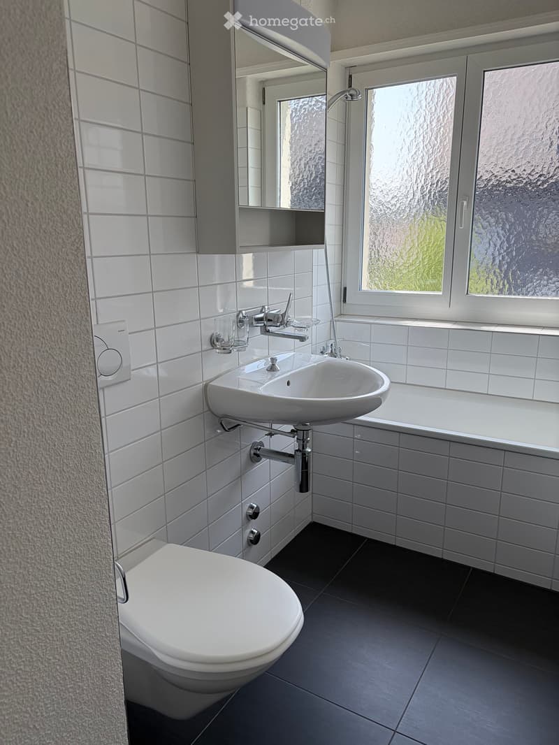 Modern bathroom with white tiles, a toilet, a sink with a modern faucet, a mirror, and a window with frosted glass.