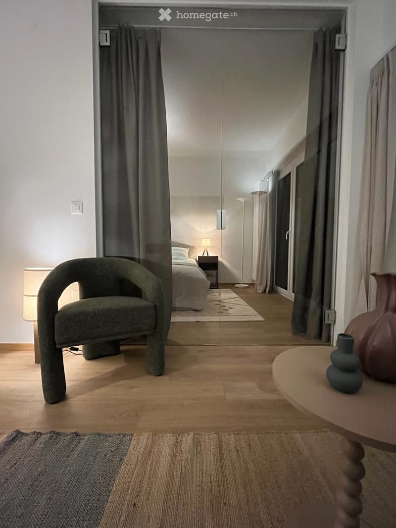 Modern living space with a green armchair, round wooden table with vases, and a visible bedroom area with a bed and nightstand.