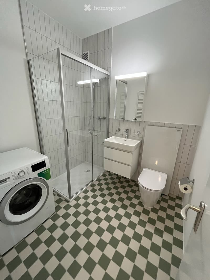 Modern bathroom with green and white checkered floor, white toilet, sink, mirror, shower, and washing machine.