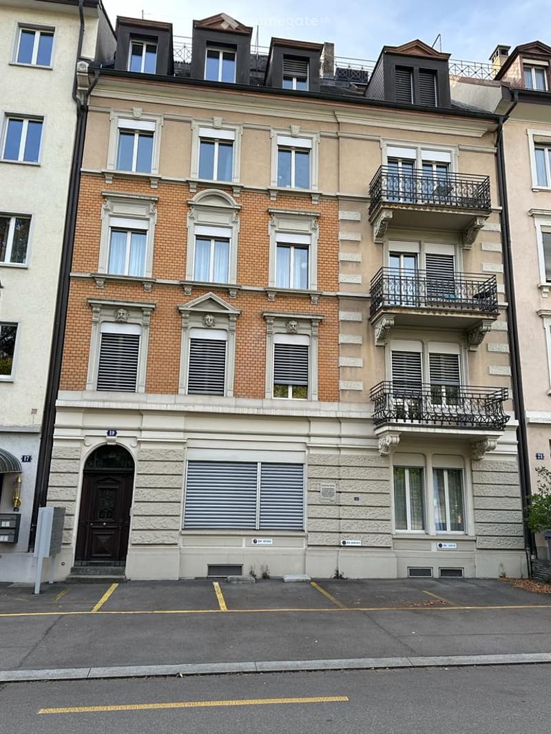 Three-story brick and concrete building with balconies, double doors, multiple windows, parking area in front