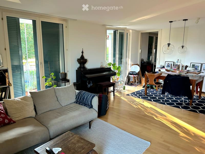 Bright living room with a grey sectional sofa, wooden coffee table, piano, dining table with chairs, and pendant lights. Large sliding glass doors lead to a balcony.