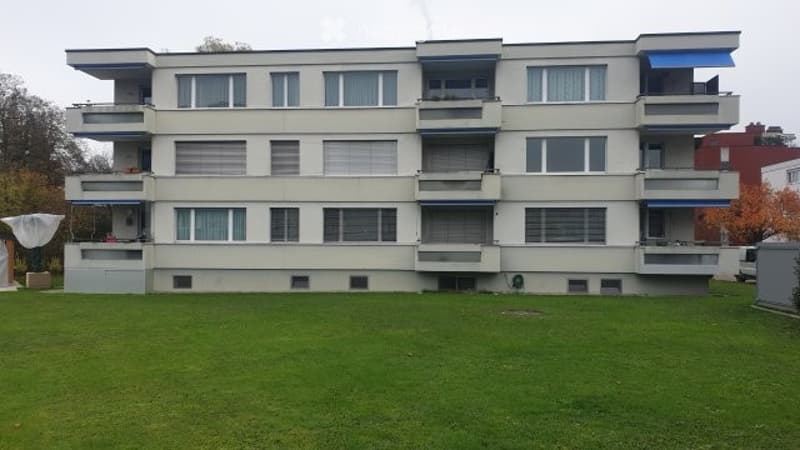 Modern multi-story apartment building with balconies and large windows, surrounded by a green lawn.