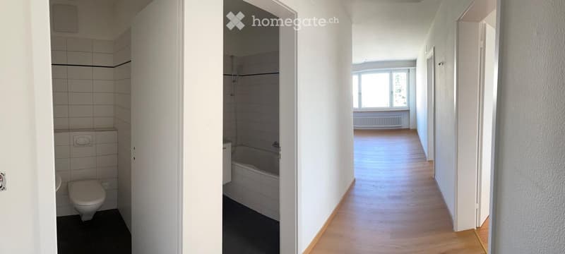 Empty hallway with wooden flooring, white walls, and doors leading to a bathroom and another room. The bathroom contains a toilet and a bathtub.