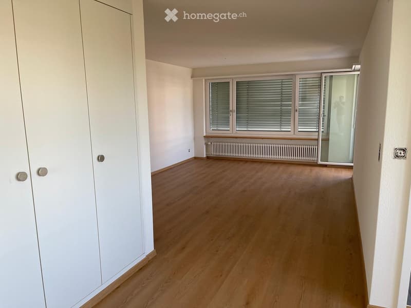 Empty room with light wooden flooring, white walls, large windows with blinds, and a radiator under the windows.