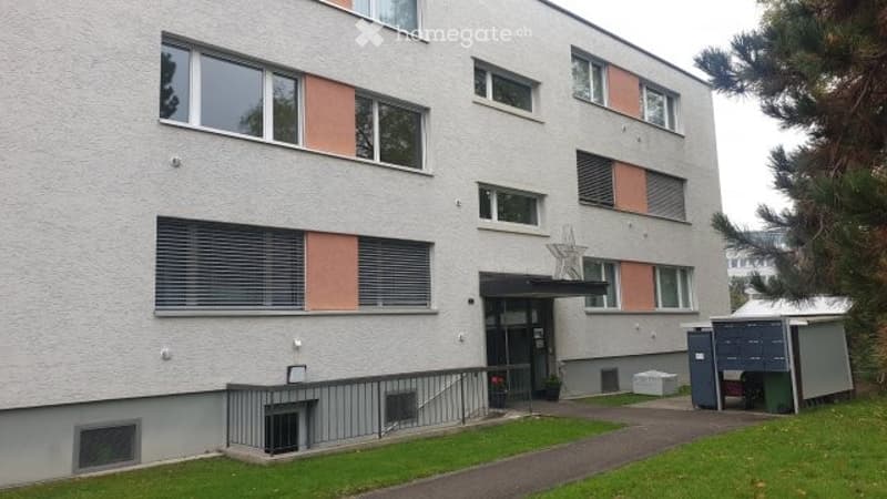 A modern multi-story apartment building with a clean, light-colored facade, large windows with blinds, and a small entrance area with a staircase leading up to the main door.