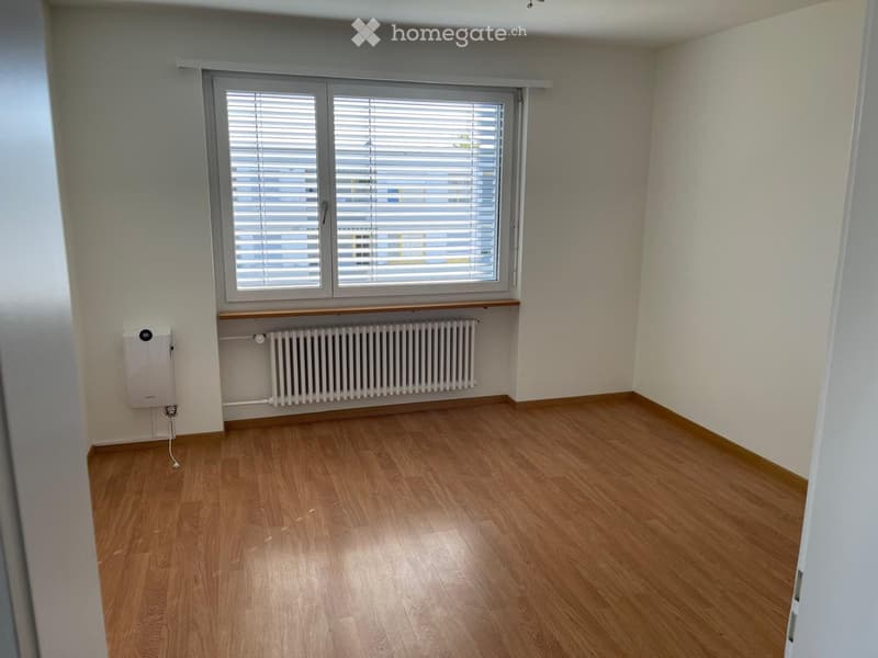 Empty room with wooden flooring, white walls, a window with blinds, a radiator, and a small shelf above it.