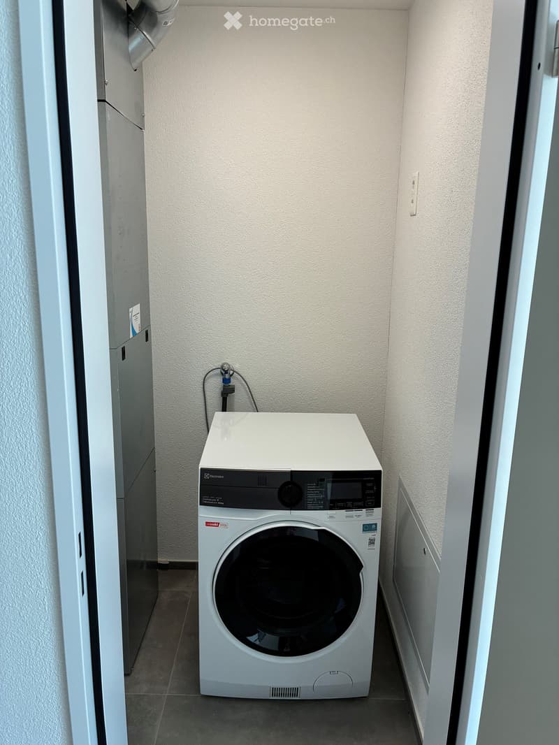 A small, modern laundry room with a white washing machine, tiled floor, and neutral-colored walls.