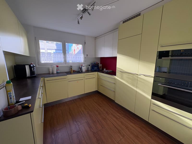 Modern kitchen with light yellow cabinets, dark countertops, and wooden flooring. Features include a coffee maker, oven, sink, and various small items on the counter.
