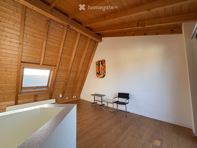 Bright, wooden-framed attic room with exposed wooden beams, a large window, a small table, a chair, and a colorful picture on the wall.
