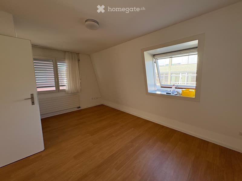 Empty attic room with wooden flooring, slanted ceilings, and two windows, one with blinds and the other open showing a view of neighboring buildings.