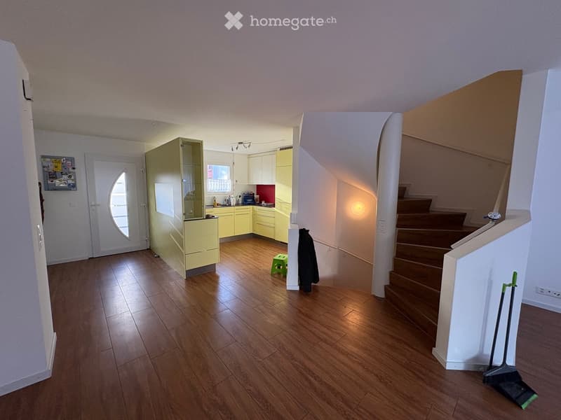 The image shows a well-lit, modern hallway with wooden flooring, leading to a small kitchen area with light yellow cabinets and a staircase. A green stool and a coat are placed near the kitchen, and a broom is leaning against the wall near the staircase.