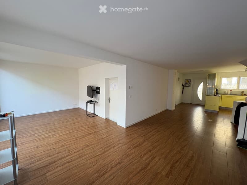 Empty room with wooden flooring, white walls, and a small TV stand in the corner. The adjacent kitchen area has yellow cabinets and a sink.