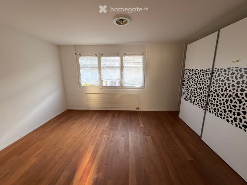 Empty room with wooden flooring, white walls, and a large window with blinds. A radiator is visible below the window, and a patterned wall design is on the right side.