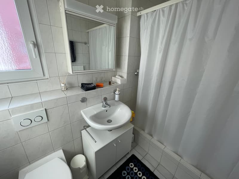 Modern bathroom with a white sink, mirror, cabinet, toilet, and a towel on the counter. The room features tiled walls and a small window with a white frame.