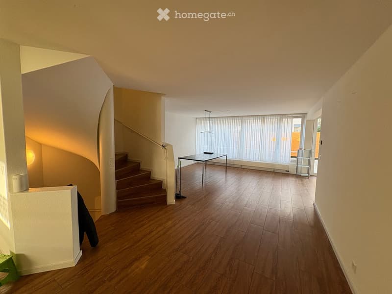 Empty hallway with wooden flooring, a staircase leading upwards, and a small table near a large window with blinds.